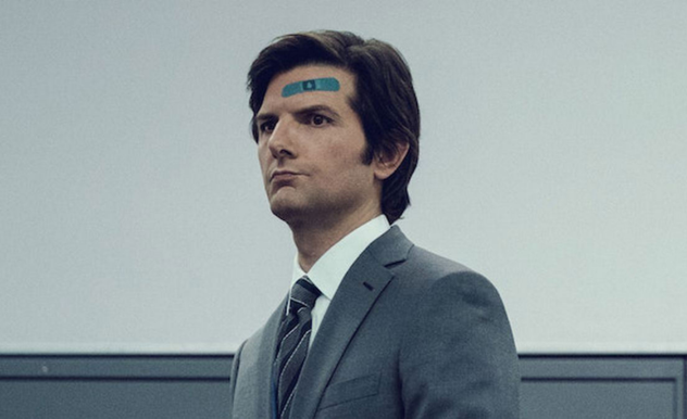 A man with quaffed, dark hair stands in a room with various shades of grey on the walls. He had a blue bandage on his forehead.