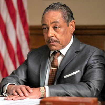 Giancarlo looking tired/annoyed in a gray suit and brown checkered tie with a gold tie pin. He is sitting behind a deck with a reddish brown book on the table with an American Flag in the background.