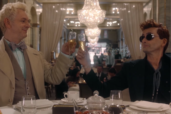 In the Ritz classy dinning area, David Tennant is wearing a black outfit and circular black glasses. Michael Sheen is wearing a tan and brown suit with a bowtie. Michael and David are clinking their glasses of bubbly wine.