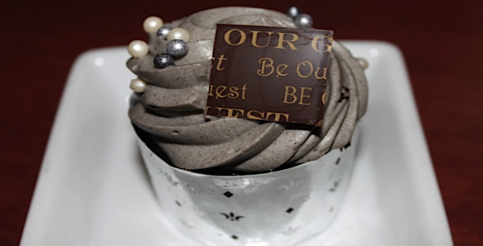 A spiral of whipped gray mousse, topped with silver, gray, and white chocolate pearls, with a silver serving bowl, and a piece of chocolate with "Be Our Guest" printed in gold, in varying fonts, on atop the mousse.