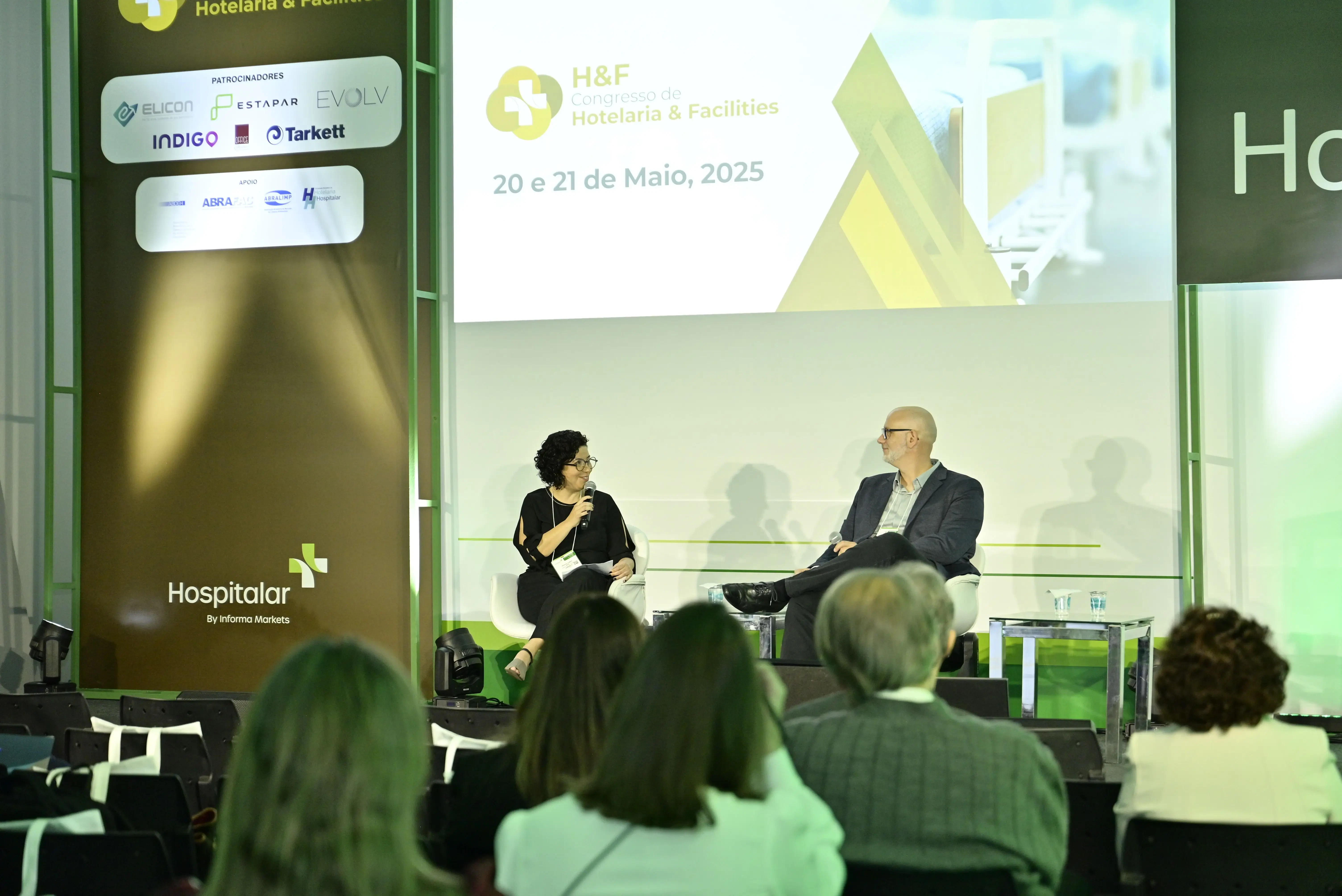 Vista frontal do palco do Congresso H&F. Uma mediadora e um palestrante estão sentados em poltronas brancas realizando uma entrevista. No telão ao fundo, o logotipo do 'H&F Congresso de Hotelaria & Facilities' e as datas do evento. À esquerda, um banner exibe as marcas patrocinadoras e o logo da Hospitalar.