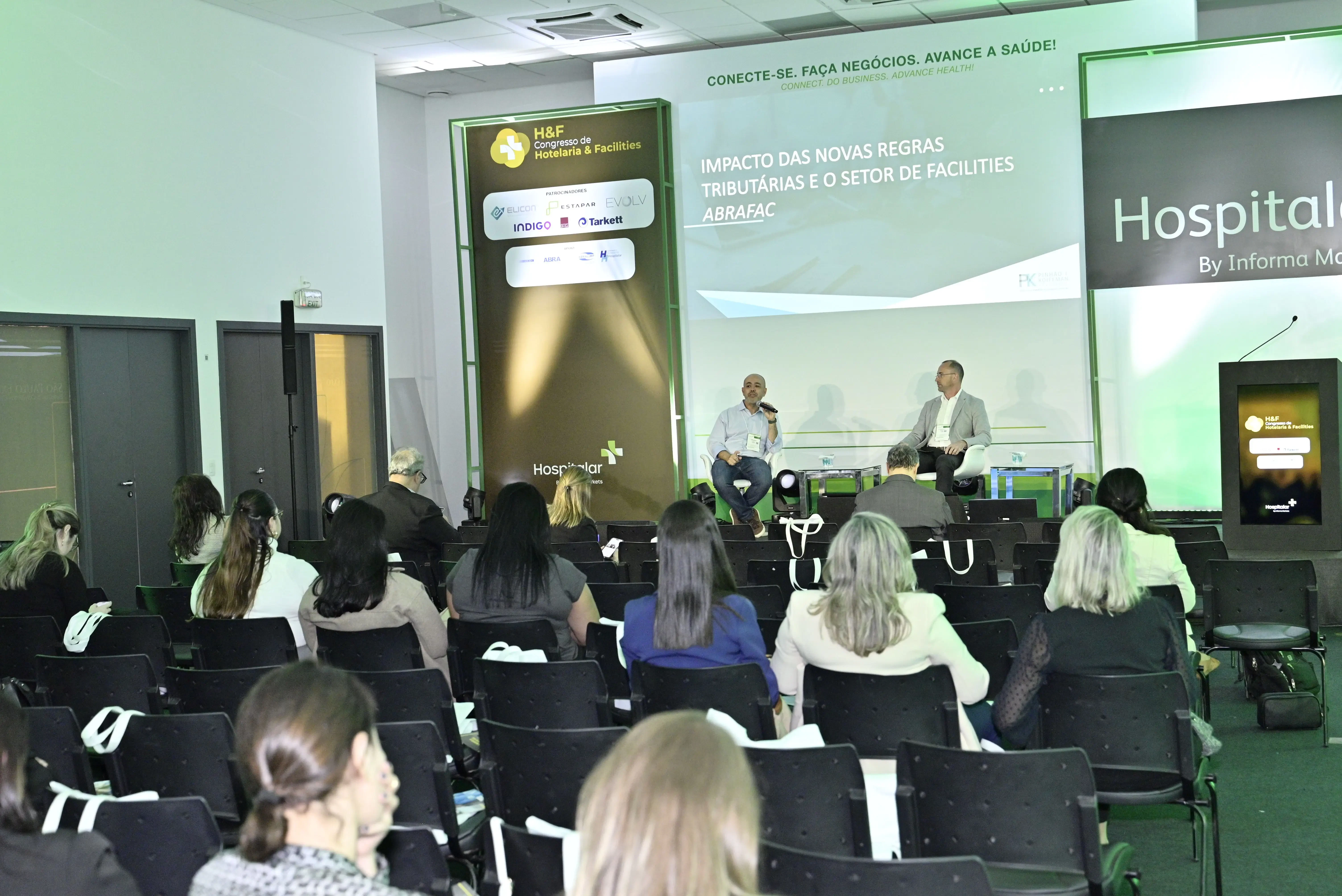 Vista da plateia em direção ao palco durante palestra sobre o 'Impacto das novas regras tributárias e o setor de Facilities'. Dois especialistas participam de um painel de discussão. O ambiente está organizado com cadeiras pretas e iluminação profissional.