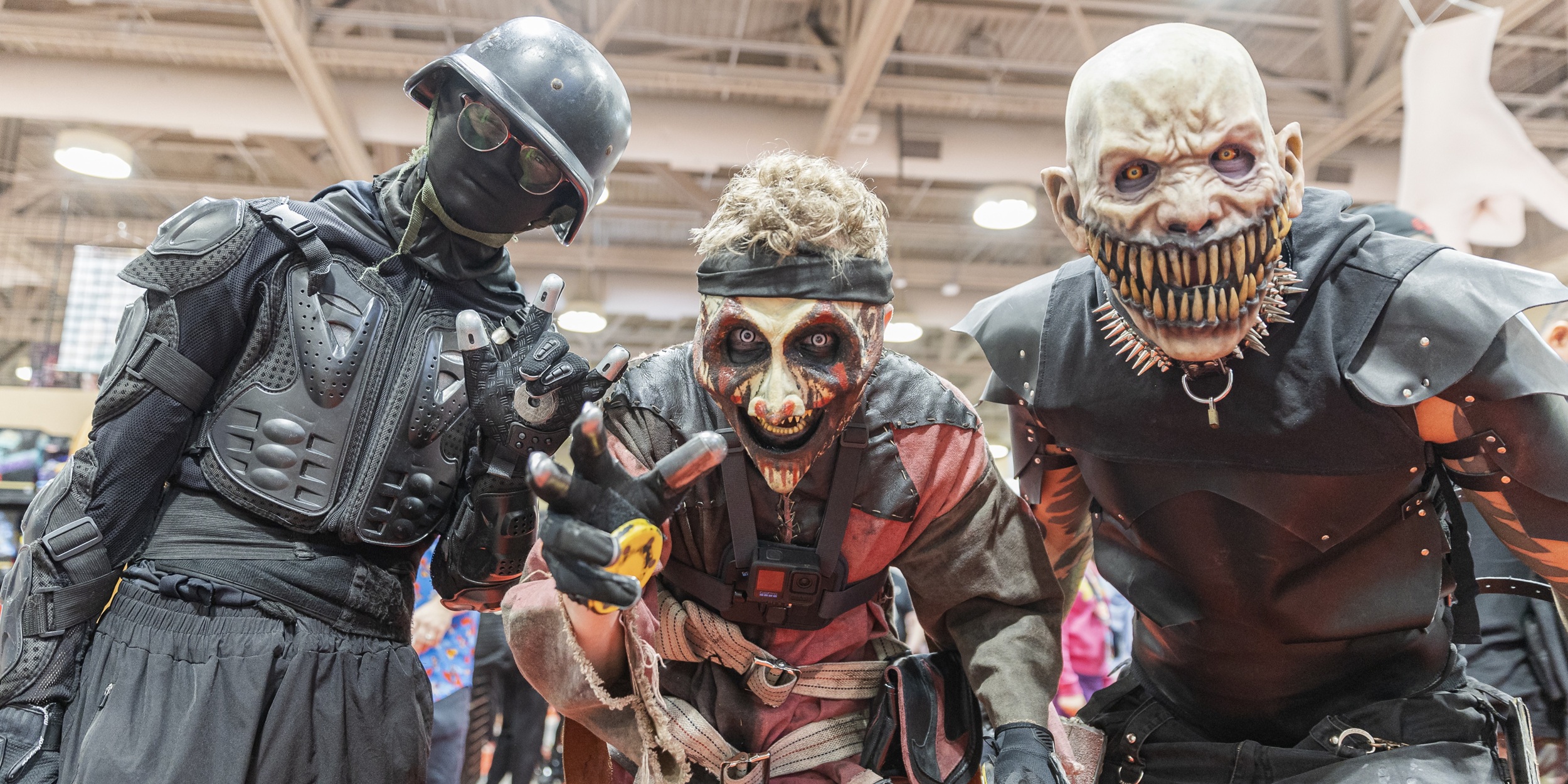 Three horror-themed cosplayers lean toward the camera on a convention floor, wearing detailed monster makeup and dark tactical-style costumes, including snarling masks and armored outfits, creating an intense and dramatic group pose.
