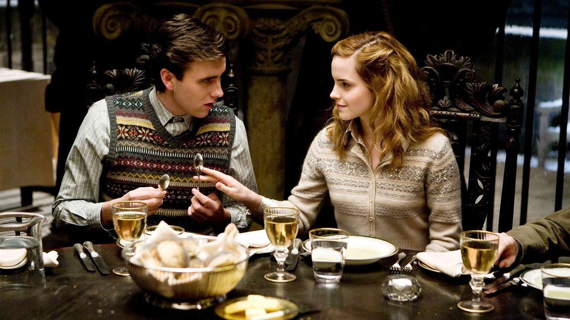 Sitting next to Hermione at a dinner table, Neville is holding two spoons while Hermione is reaching out to touch one of the spoons. They are making eye contact.