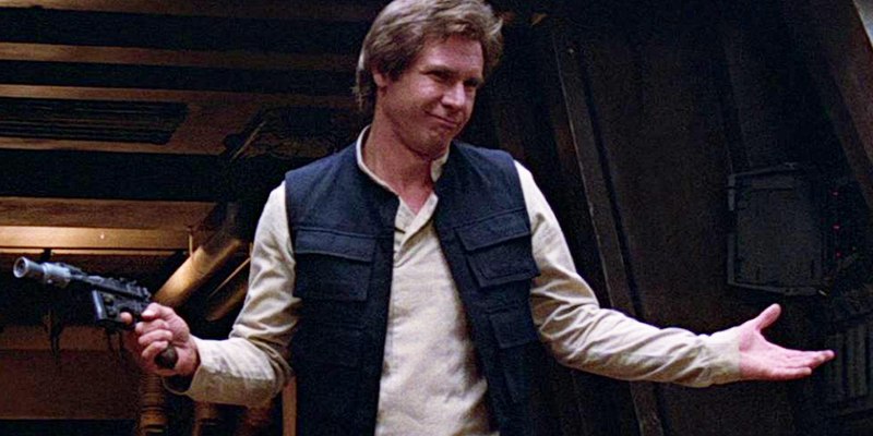 Han Solo shrugs, he holds his blaster with a finger on the trigger. He half smiles as he listens to the person speaking off screen.