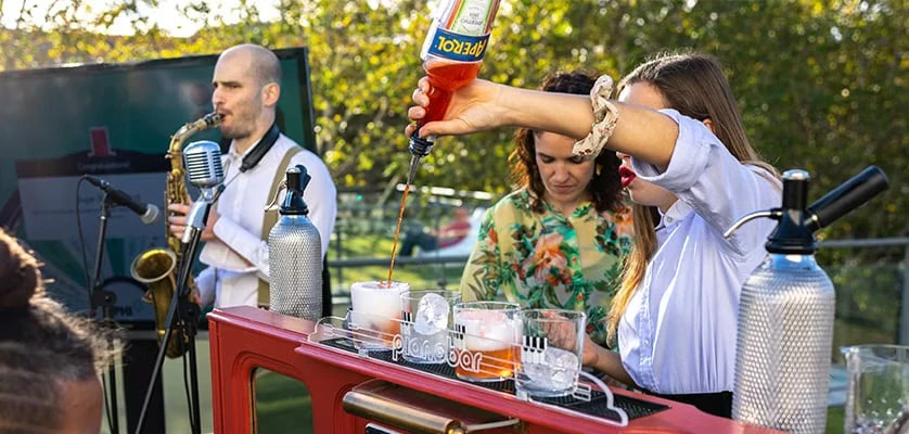 Corso Italia happy hour at CPHI Milan showing a bar tender pouring an aperol spritz and a musican playing a saxophone behind a bar