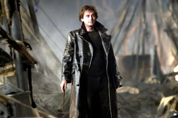 In a long leather trench coat and messy hair, David Tennant holds a wand and is standing a disaster scene