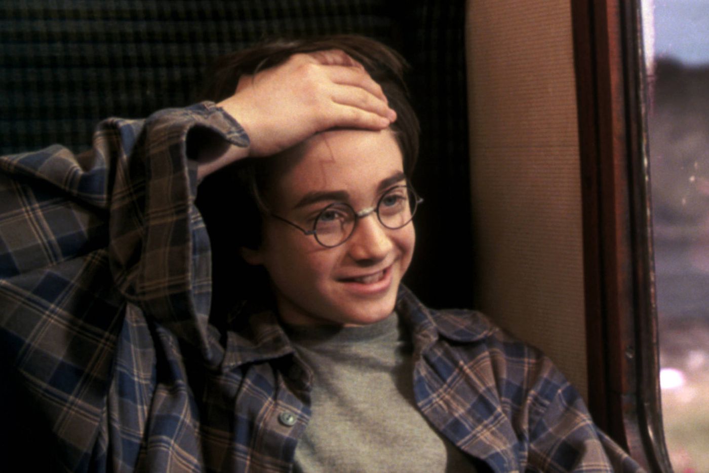 Harry Potter holds up his hair with one hand to expose the scar on his forehead. He is wearing a grey shirt with a plaid button up shirt over top. He is sitting on a train