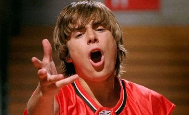 Zac Efron as Troy Bolton in a red jersey singing with his arm extended to the camera