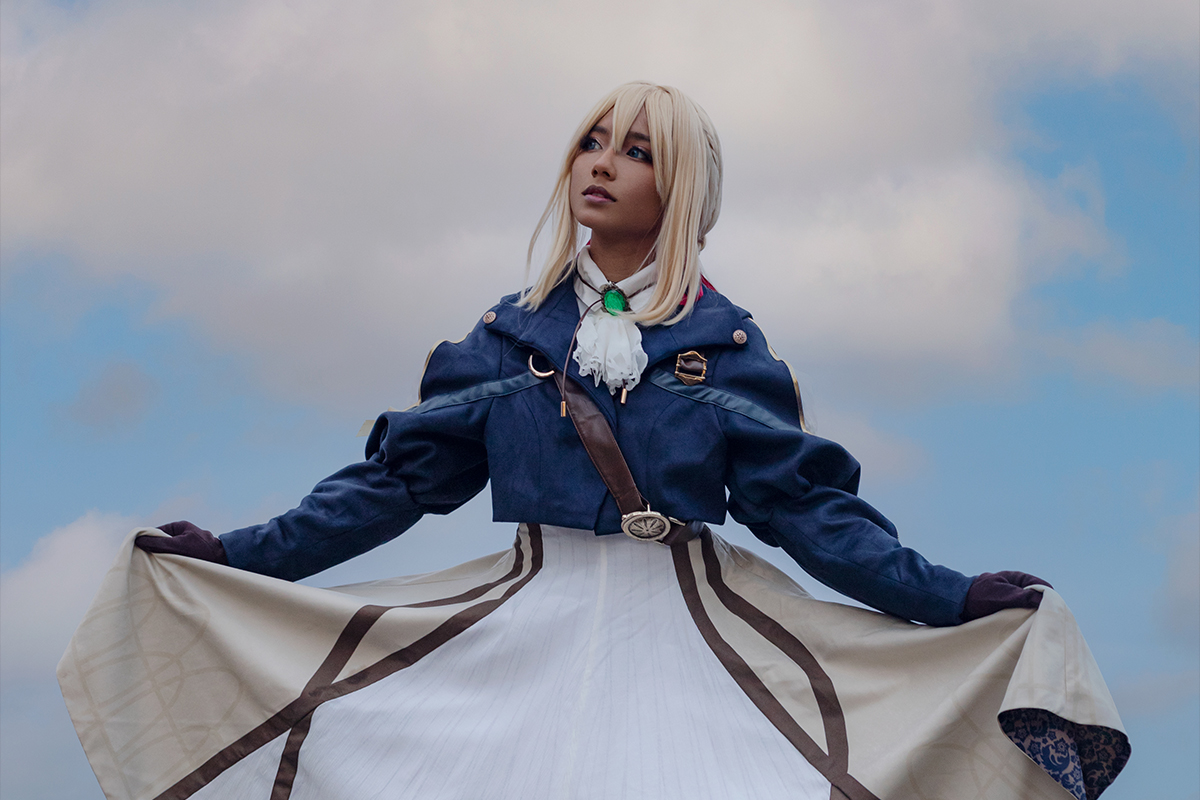 Cosplayer poses in a long brown dress with a blue short jacket over top. There are cloudy skies in the background..