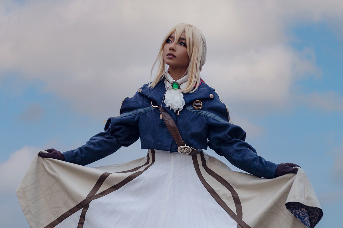 Cosplayer poses in a long brown dress with a blue short jacket over top. There are cloudy skies in the background..