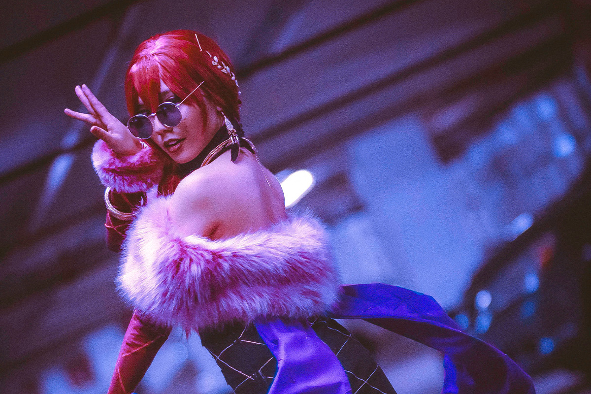 Cosplayer poses in a dress with a pink fur lining. She is wearing a bright pink wig and round sunglasses.