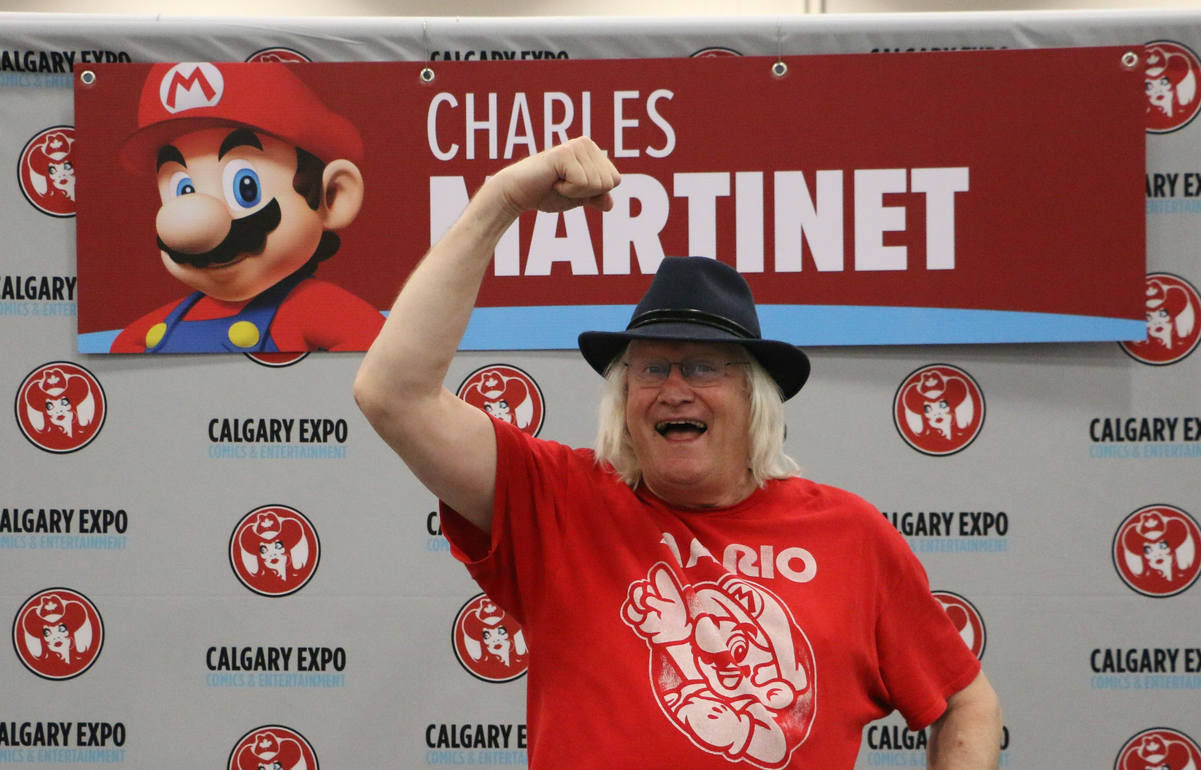 Charles Martinet in a black fedora and a red Mario shirt. He is posing in the Mario jump pose in front of his autograph table.