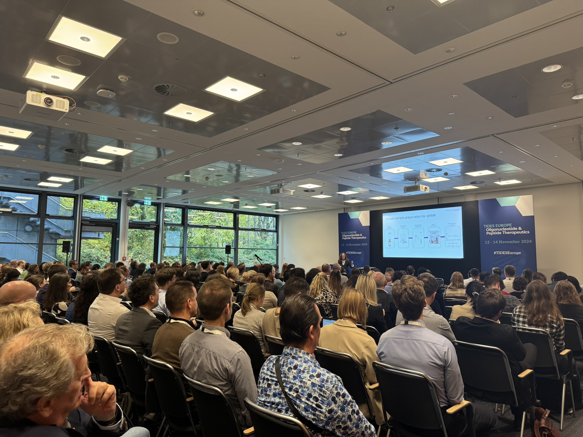 Attendees sit in a large room watching a presenter discuss a slide projected on a screen at the front of the room