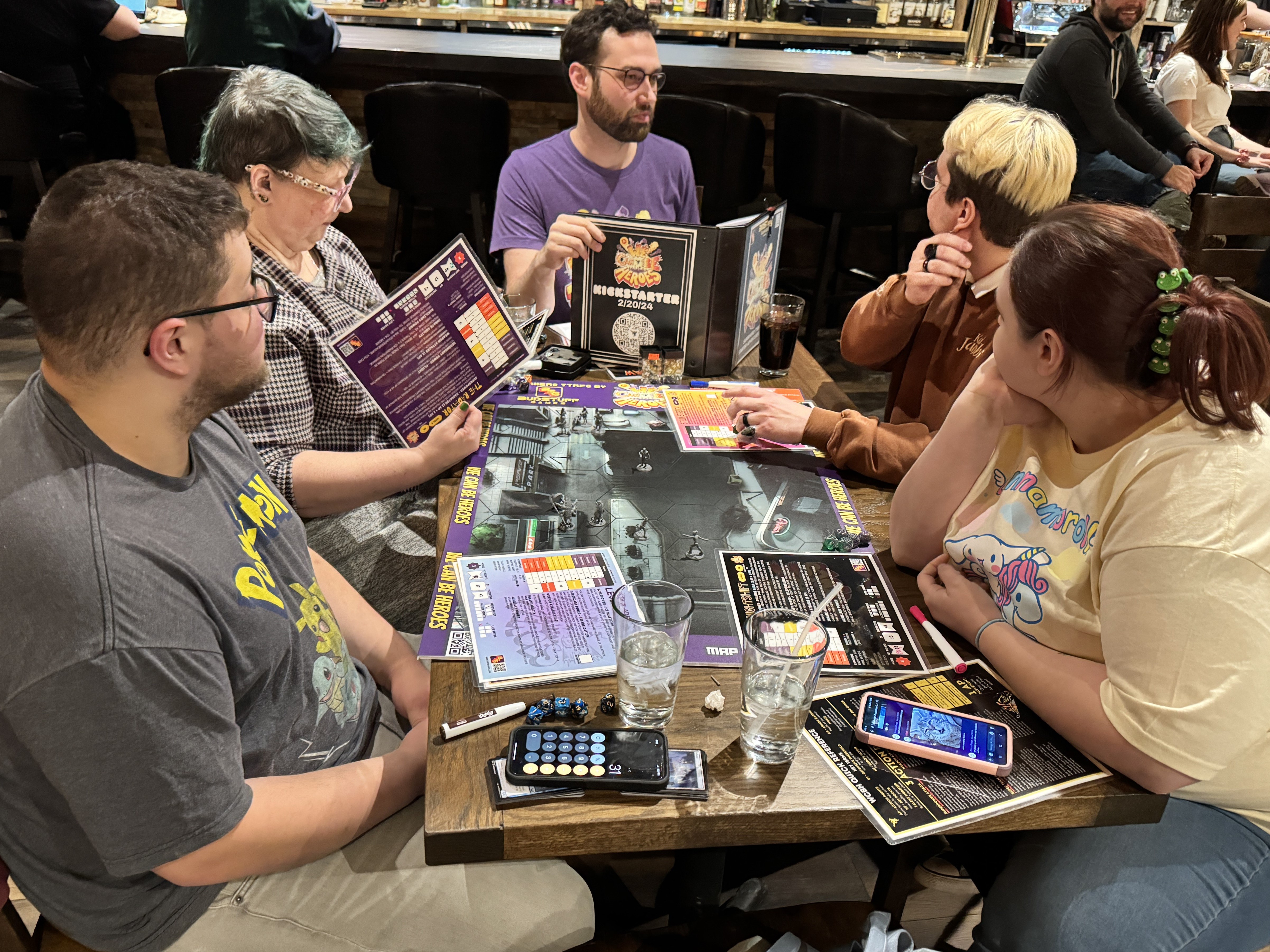 A group of people sitting around a table at a board game night, engaging with game materials and chatting. The table is filled with game boards, rule books, and dice, while the atmosphere appears friendly and focused.