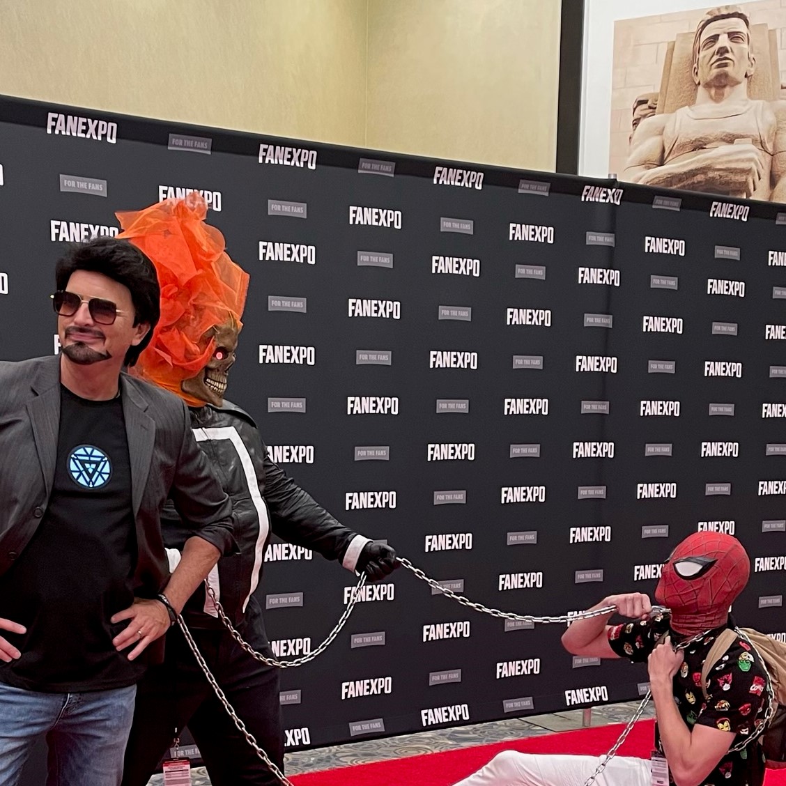 Image from the red carpet. Tony Stark, in a gray suit jacket black shirt with his reactor shining through and blue jeans, is looking away from the tom foolery of Ghost Rider, movie accurate cosplay with a flaming skull head, holding chains around a "Spiderman at a con" who is kneeling down. The Spiderman is holding the chains that are looped around the back of his neck (the chain is draped behind his neck like a large necklace) and is wearing a casual white pants, marvel button up with heads of avengers and a brown backpack. In the background, is the FAN EXPO background that repeats FAN EXPO.