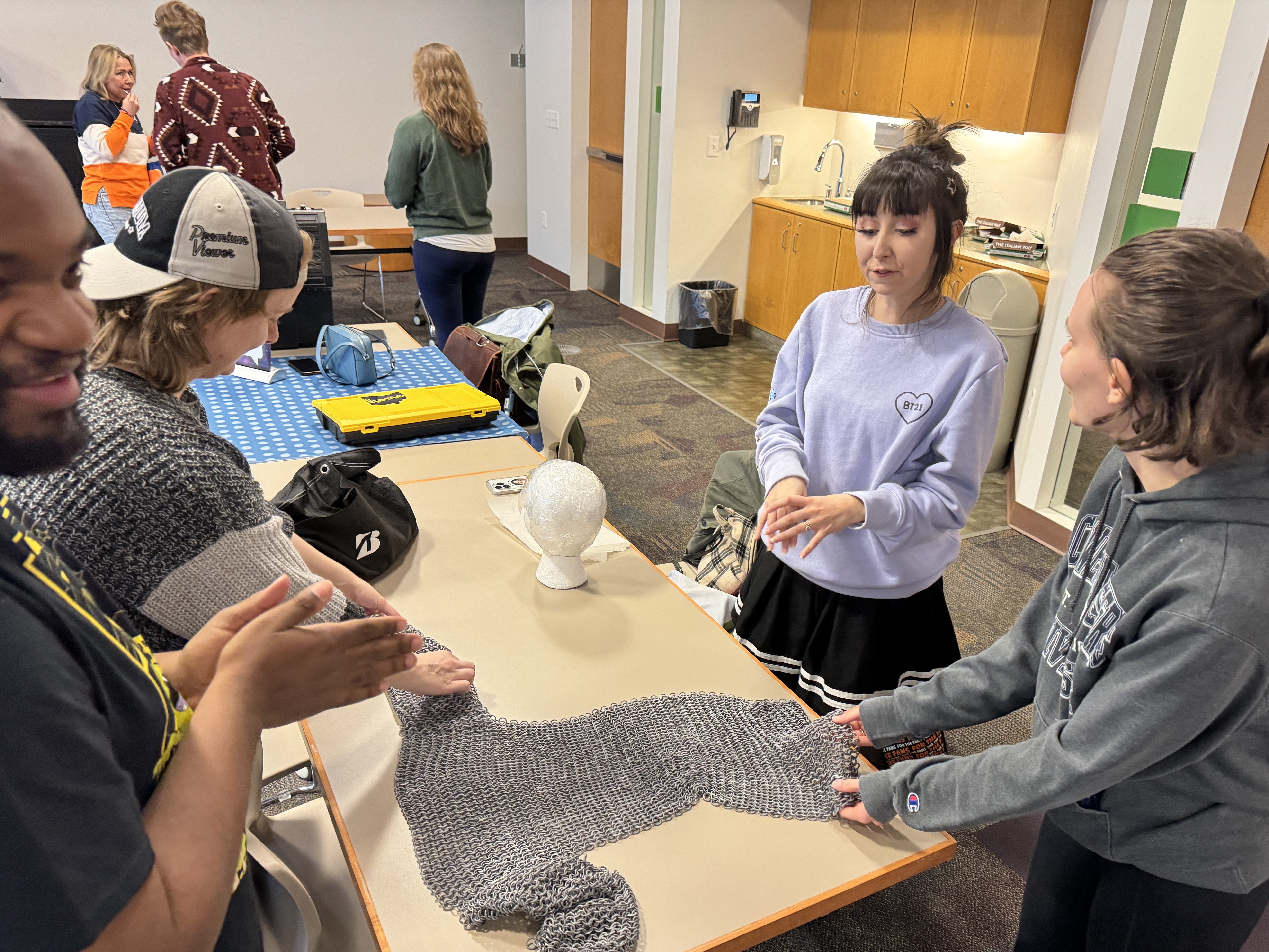 A group of people gathered around a table, discussing chainmail crafting techniques. One person holds up a piece of chainmail, while others observe and listen attentively in a well-lit classroom or workshop space.