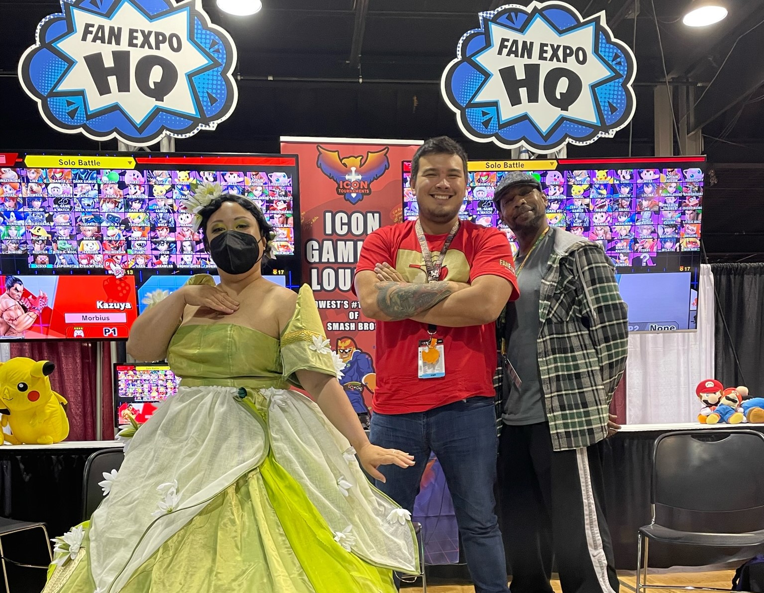 3 people standing on a stage with the FAN EXPO HQ Gaming zone with 2 monitors behind them. The person to the left is cosplaying as Tiana, the middle is wearing a red FAN EXPO Chicago 50th shirt, and the one on the right is wearing a green plaid jacket and a newsboy hat.