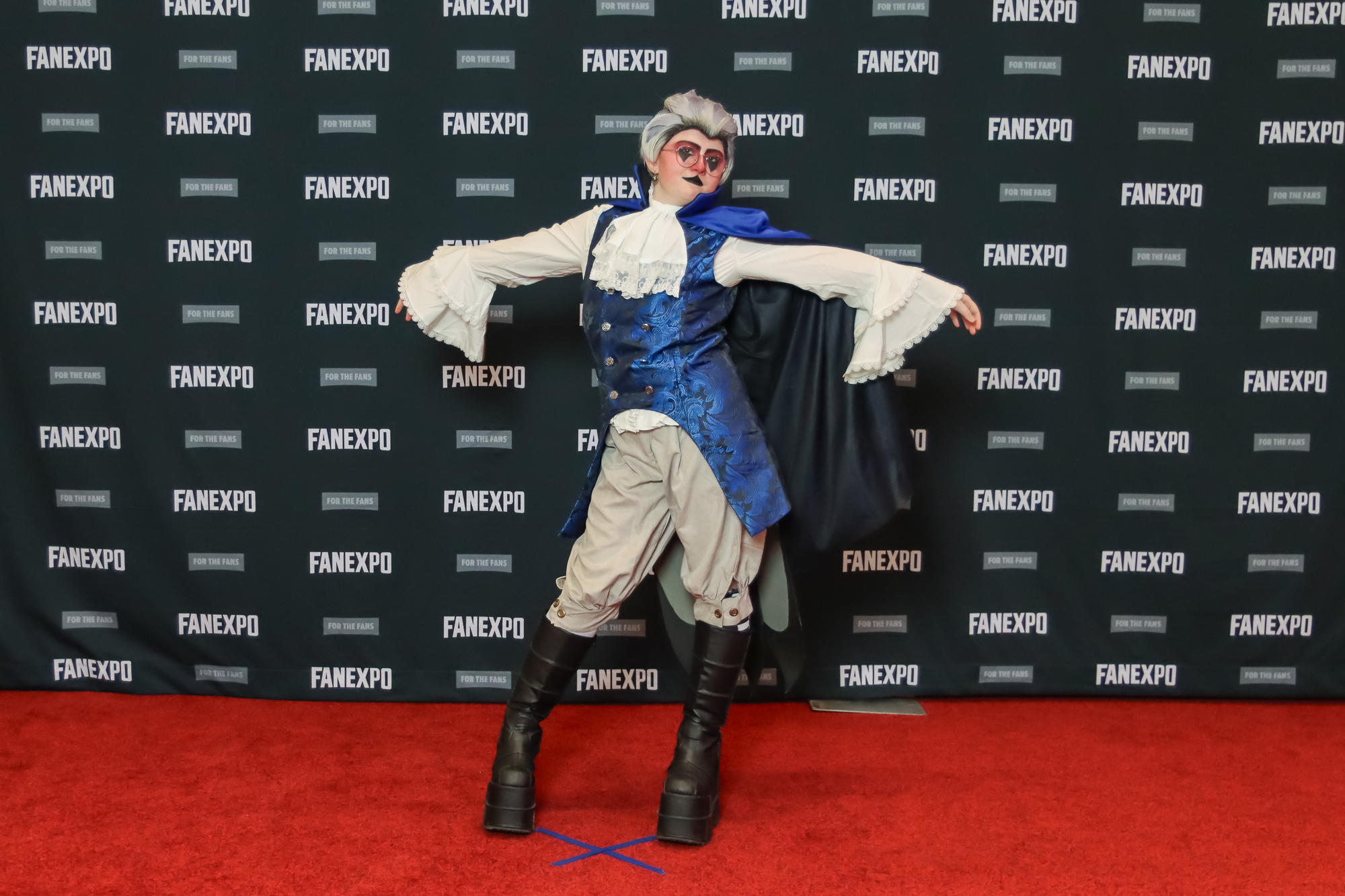 The image features an individual in a cosplay posing on a red carpet at what appears to be a cosplay event, as indicated by the "FAN EXPO" backdrop. Person is wearing a long blue vest over a ruffled white shirt and off-white pants tucked into knee-high black boots. They are also wearing a blue cape and have grey slicked back hair.