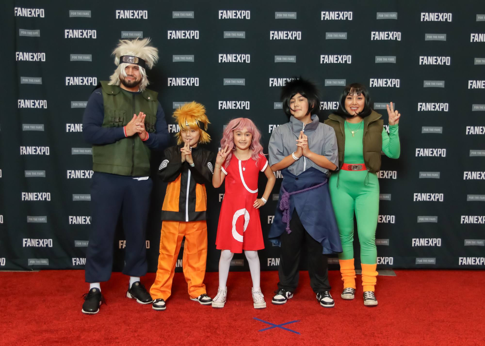 Group of 5 cosplayers standing together dressed from the anime Naruto. First person on the right is dressed as Kakashi with a green vest, black long shirt and pants with white spikey hair. Second from the right is dressed as Naruto with a orange and black tracksuit and blond spikey hair. Person in the middle is dressed as Sakura in a knee-length red dress with long pink hair. The second person from the left is dressed as Sasuke with a grey top, black pants and a dark blue over shirt with purple belt and black spikey hair. Person on the far right is dressed as Rock Lee and is wearing a green jumpsuit with orange leggings and has shoulder-length black hair.