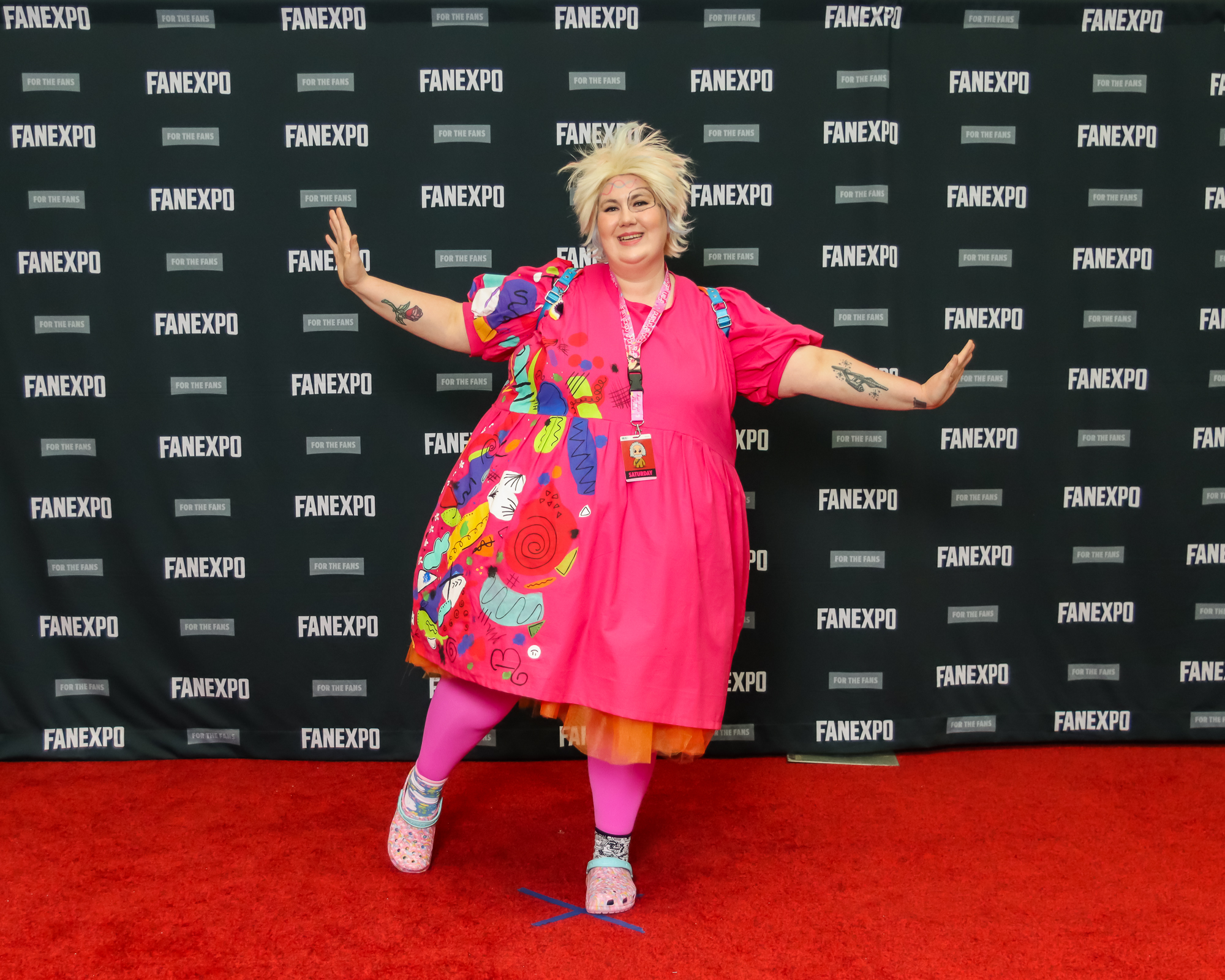 The image depicts a person striking a pose on a red carpet against a backdrop with "FAN EXPO" branding, indicative of a fan convention. Person is dressed as Weird Barbie and has chopped, short blond hair sticking up at odd angles, and is wearing a pink dress with colorful cut outs  sewn on top. They are also wearing pink tights and pink crocs.