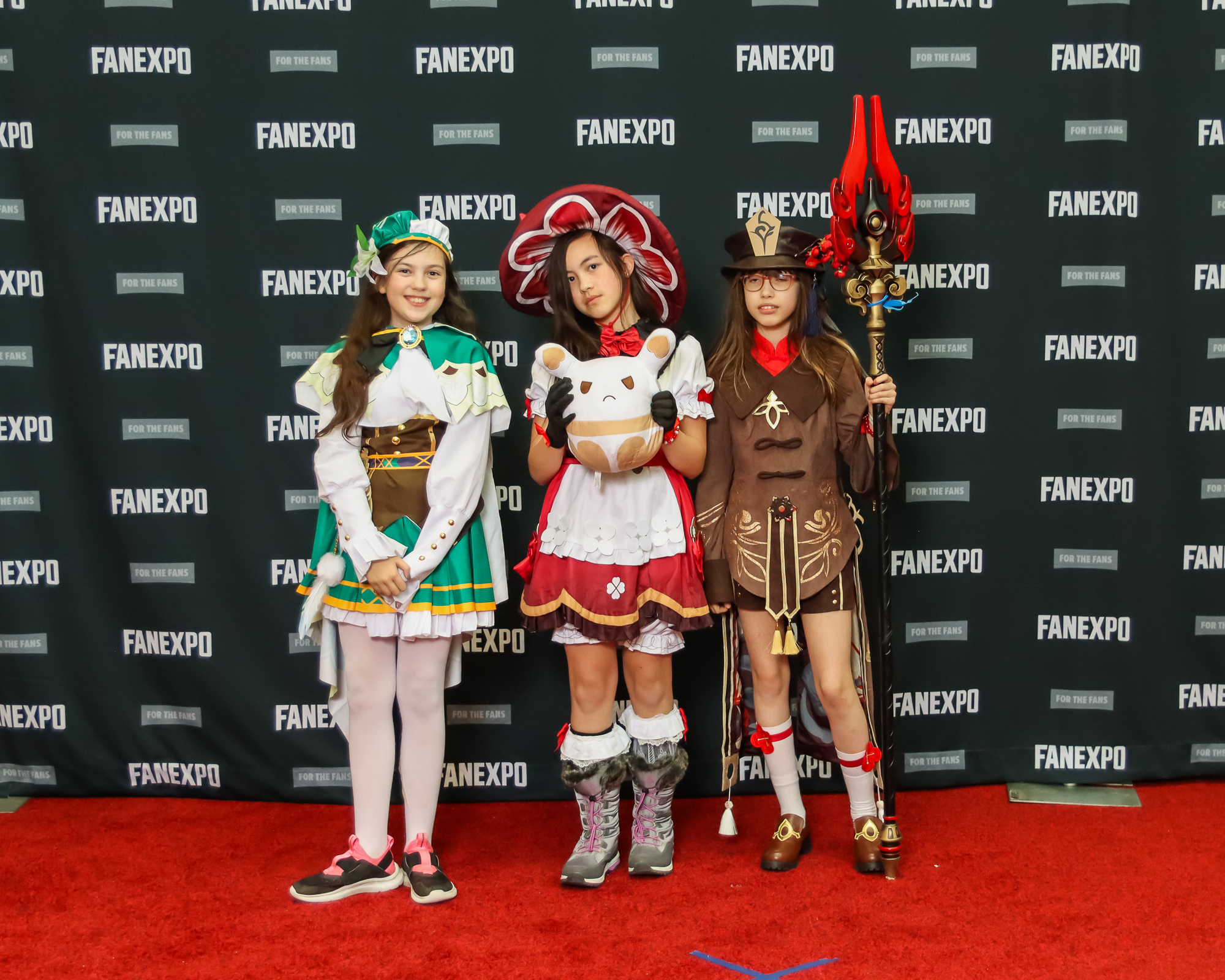 The image captures three individuals on a red carpet, each dressed in various costumes from the Genshin Impact video game. The person on the left is dressed as the character Venti with a green and white hat, and white shirt with green cape fastened with a green jewel. They have a green skirt with a large brown best around their middle and white tights. The person in the middle is dressed as the character Klee with a large circular red and white hat and a red dress with white bell sleeves and apron. The person on the right is dressed as the character Hu Tao and is wearing a black top hat, a brown overcoat with gold embellishments and brown shorts with white socks. The person also carries a pole arm with a red flame pattern.