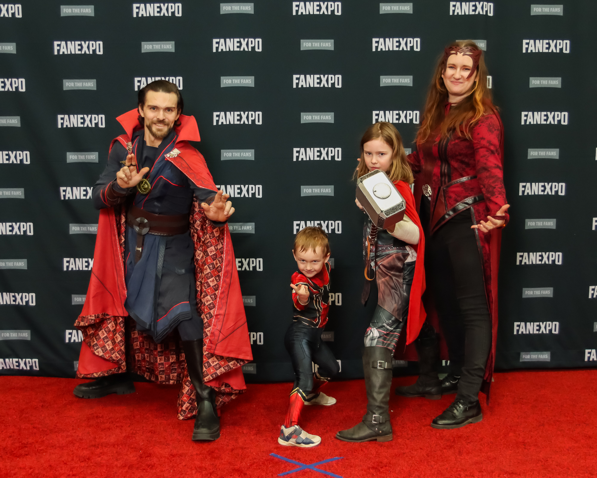 The image shows four individuals in costumes from the MCU. From left to right, the first individual is wearing Doctor Strange costume with blue robes and a red cape. The second a child wearing the black and red Spider-man suit. The third person is dressed as Thor with a red cape, brown boots and is posing with a large hammer. The fourth dressed as the Scarlet Witch and wears a red head piece, red shirt with cape and black pants.
