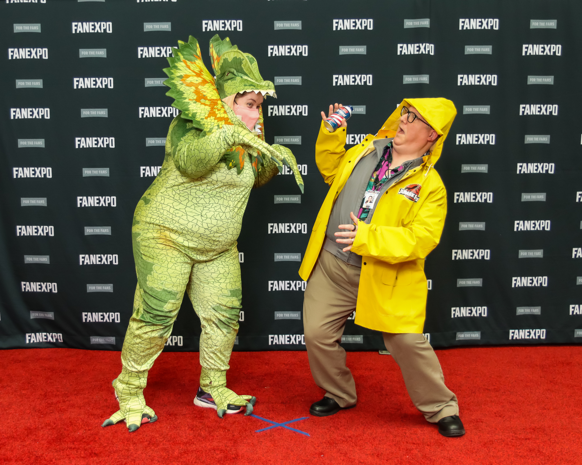 The image shows two individuals on a red carpet striking poses, dressed up from the original Jurassic Park movie. The person on the left is dressed in a full-bodied dinosaur suit which is light green and has and frilled neck collar. They are posed as if they are going to attack the person on the right. The person on the right is dressed in a yellow rain coat with glasses, a grey shirt, and tan pants who is leaning away from the dinosaur cosplayer in fear.