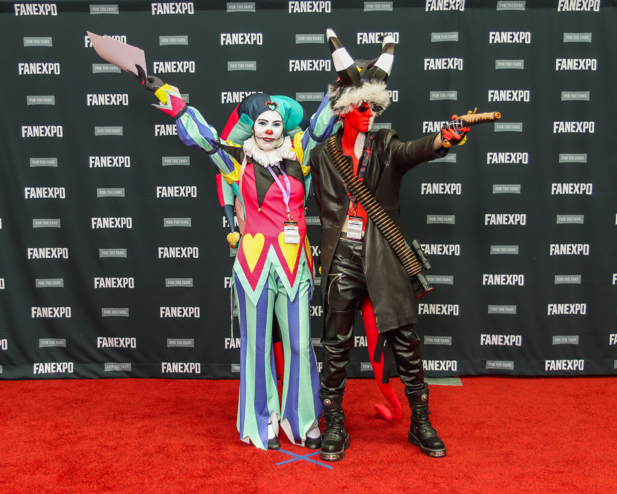 The image depicts two individuals dressed in Helluva Boss costumes, posing together on a red carpet in front of a backdrop that features the "FAN EXPO" logo. The person on the left is dressed as Fizzarolli in a colorful clown costume with red nose and two pointed hat. The person on the right is dressed as Blitzo with black and white horns on top of their head, red and white body paint, black sunglasses, a long black coat left open and black leather pants and boots. An ammo belt is wrapped across their torso and they are posing holding a gun.