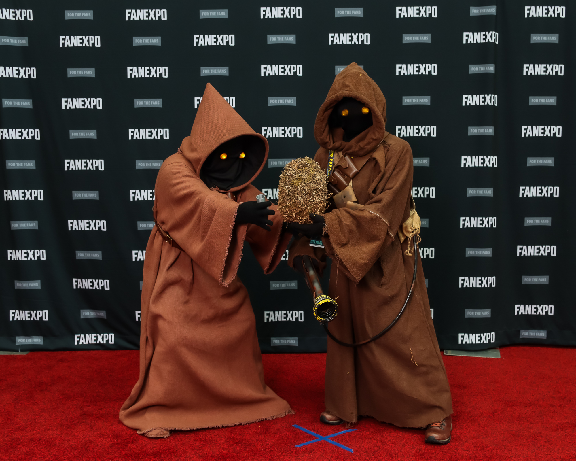The image features two individuals posing on a red carpet. They are dressed as Jawas from the Star Wars franchise and are wearing full-bodied brown cloaks with their faces hidden and glowing yellow eyes peering from their hoods.