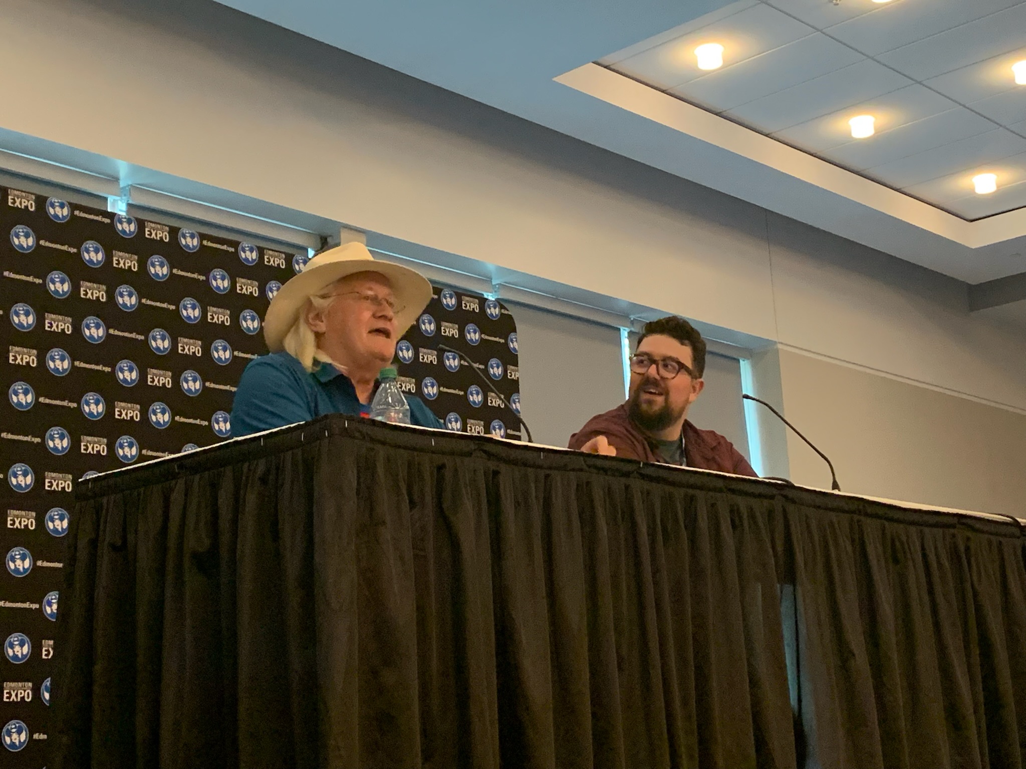 Charles Martinet in a white cowboy hat and a blue shirt with a microphone in front of him as he sits next to a bearded gentleman.