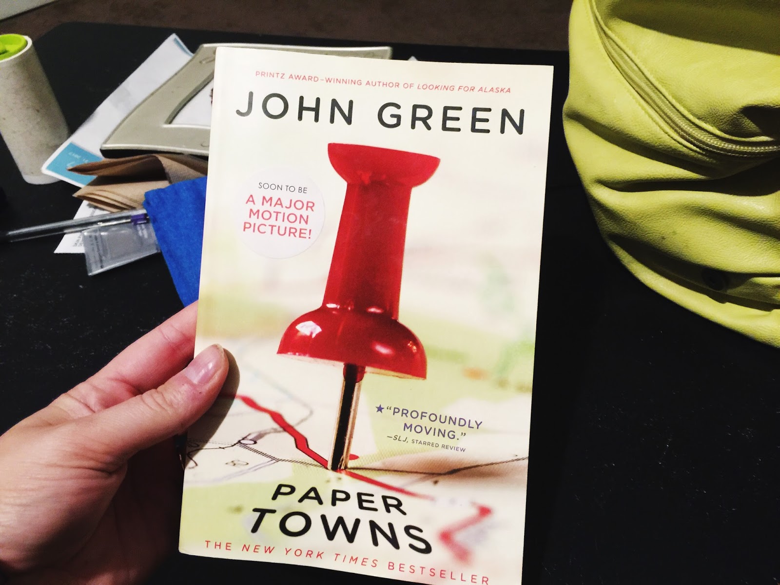 If the foreground, a person holds a paperback copy of John Green's Paper Towns. If the back, a desk with miscellaneous items stacked in a pile.