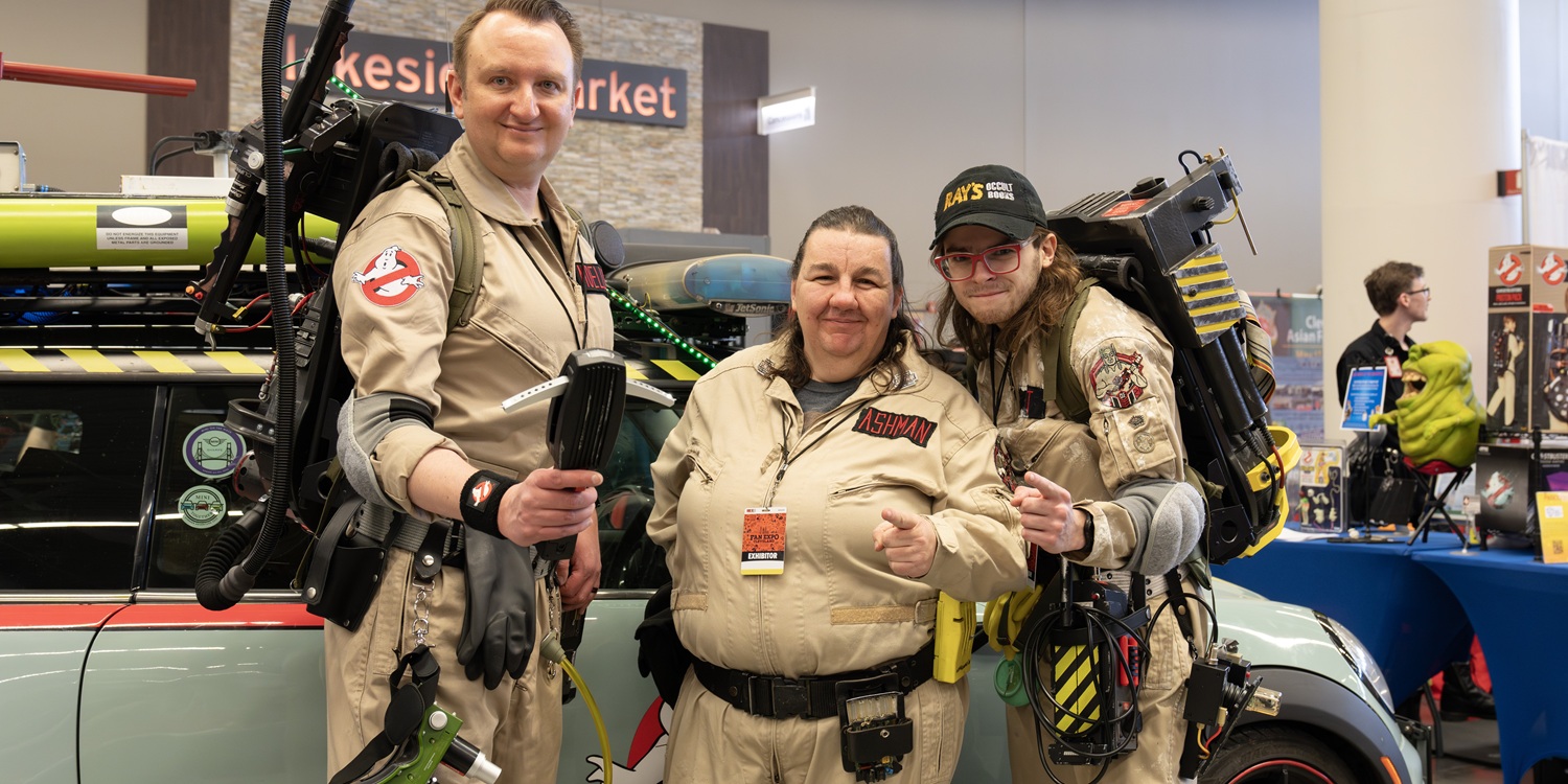 Three attendees pose together in Ghostbusters-inspired jumpsuits beside a themed display. Equipment props and signage appear behind them during an interactive paranormal experience at FAN EXPO Cleveland.