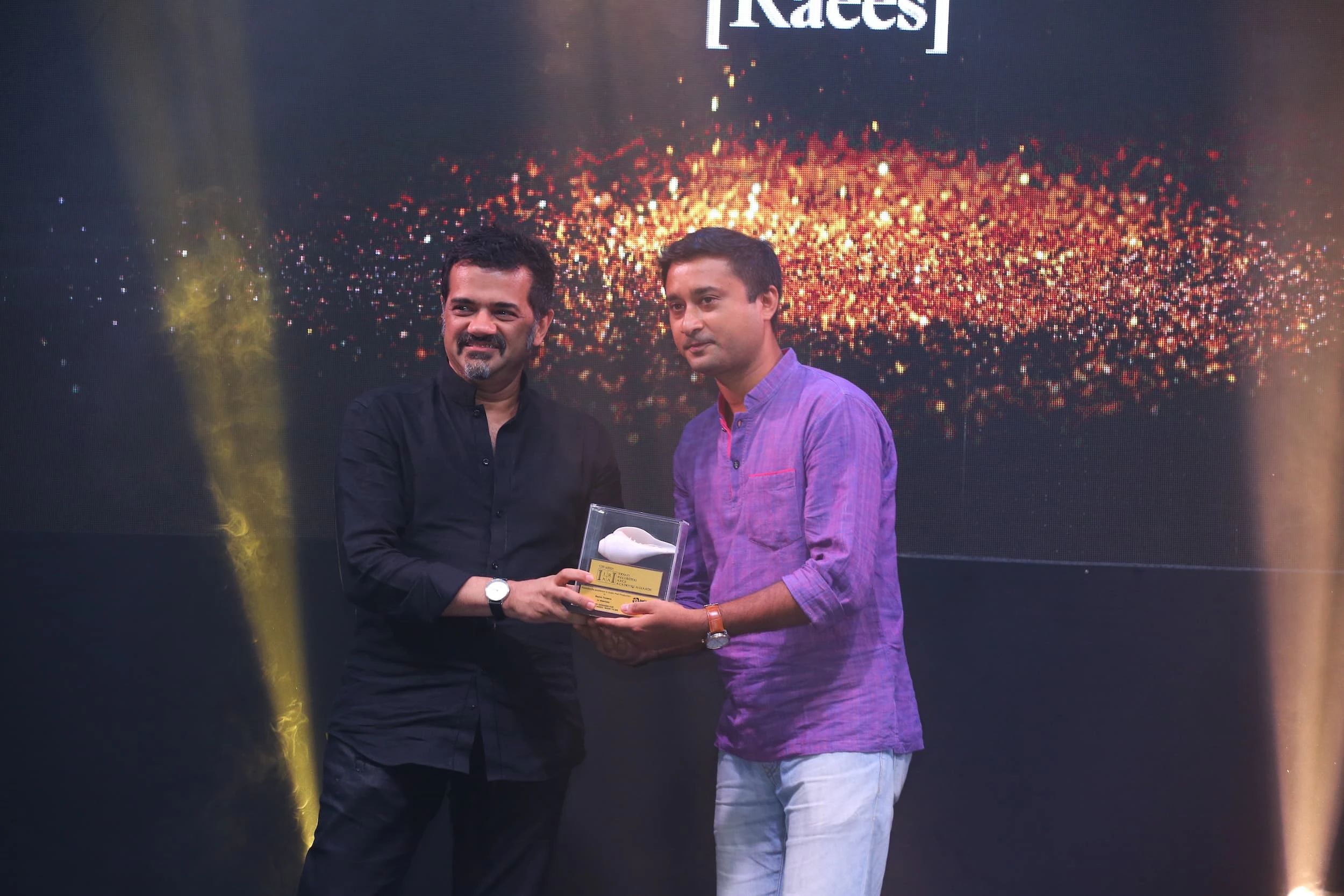 Two men posing with a trophy at the IRAA Palm Awards photo call, one dressed in black and the other in white trousers with a violet shirt, with sparkling lights creating a celebratory atmosphere.