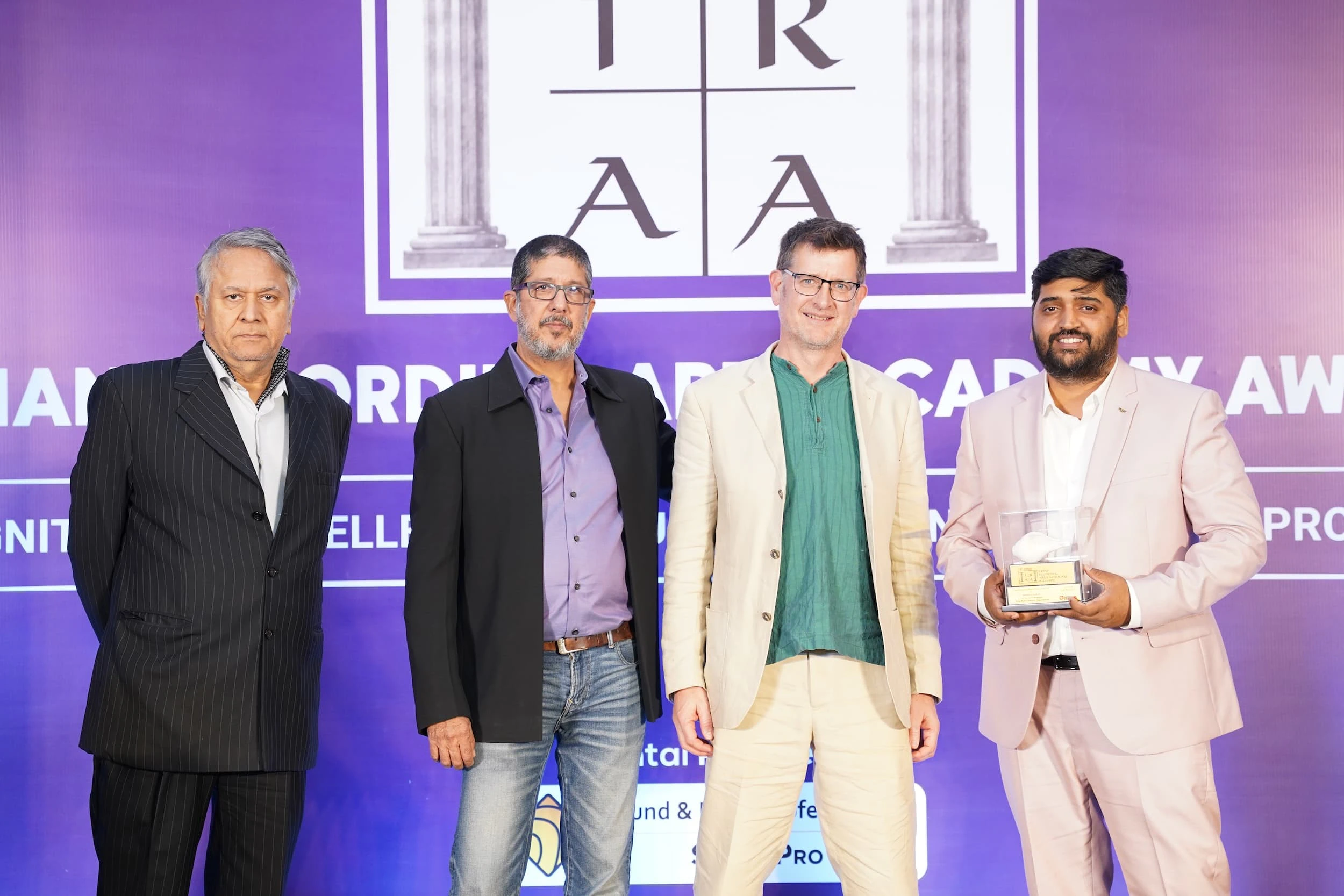 Four people posing at the IRAA Palm Awards photo call with the event logo behind them, the person on the far left holding an award.