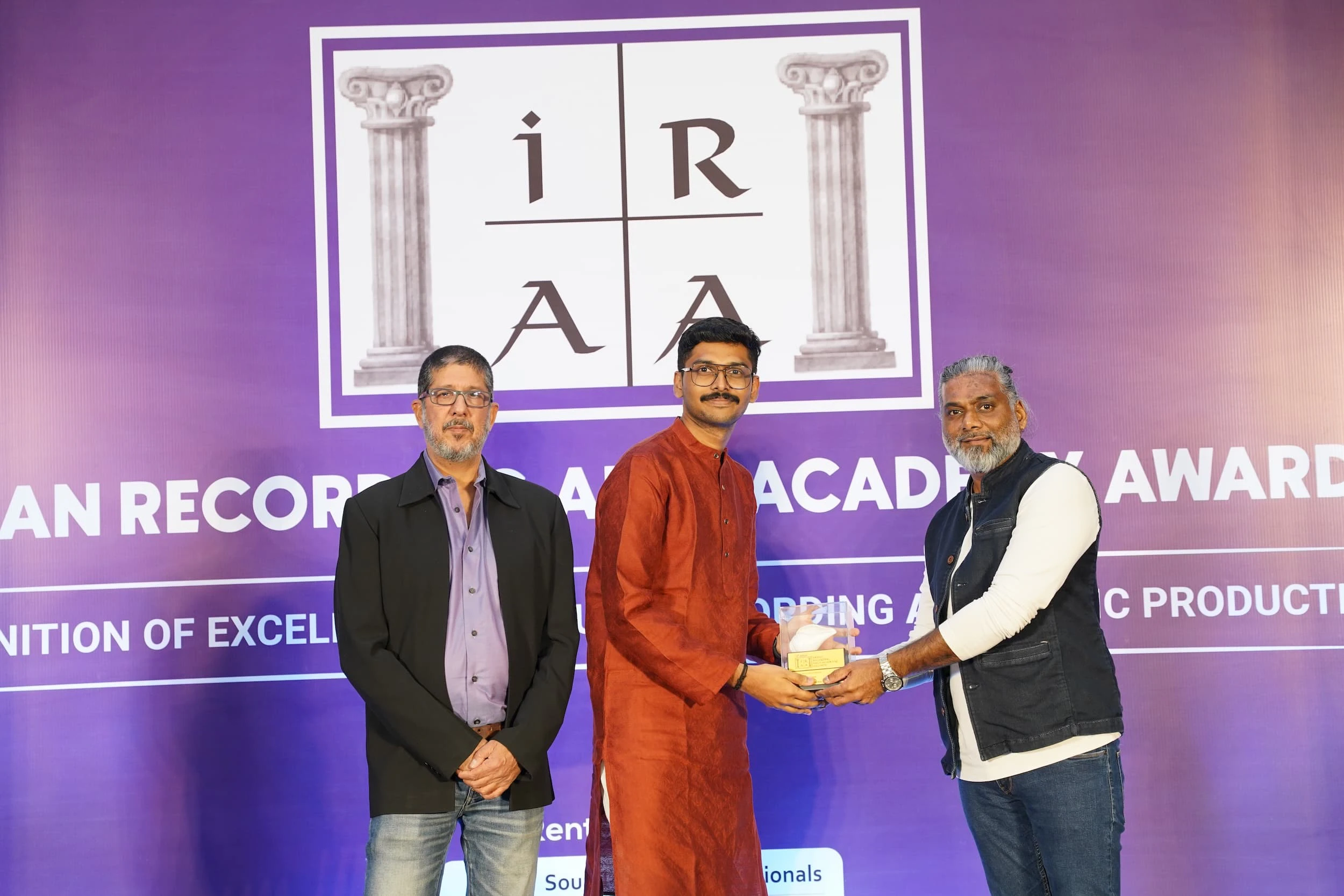 Three men at the IRAA Palm Awards, with the central person receiving a trophy.