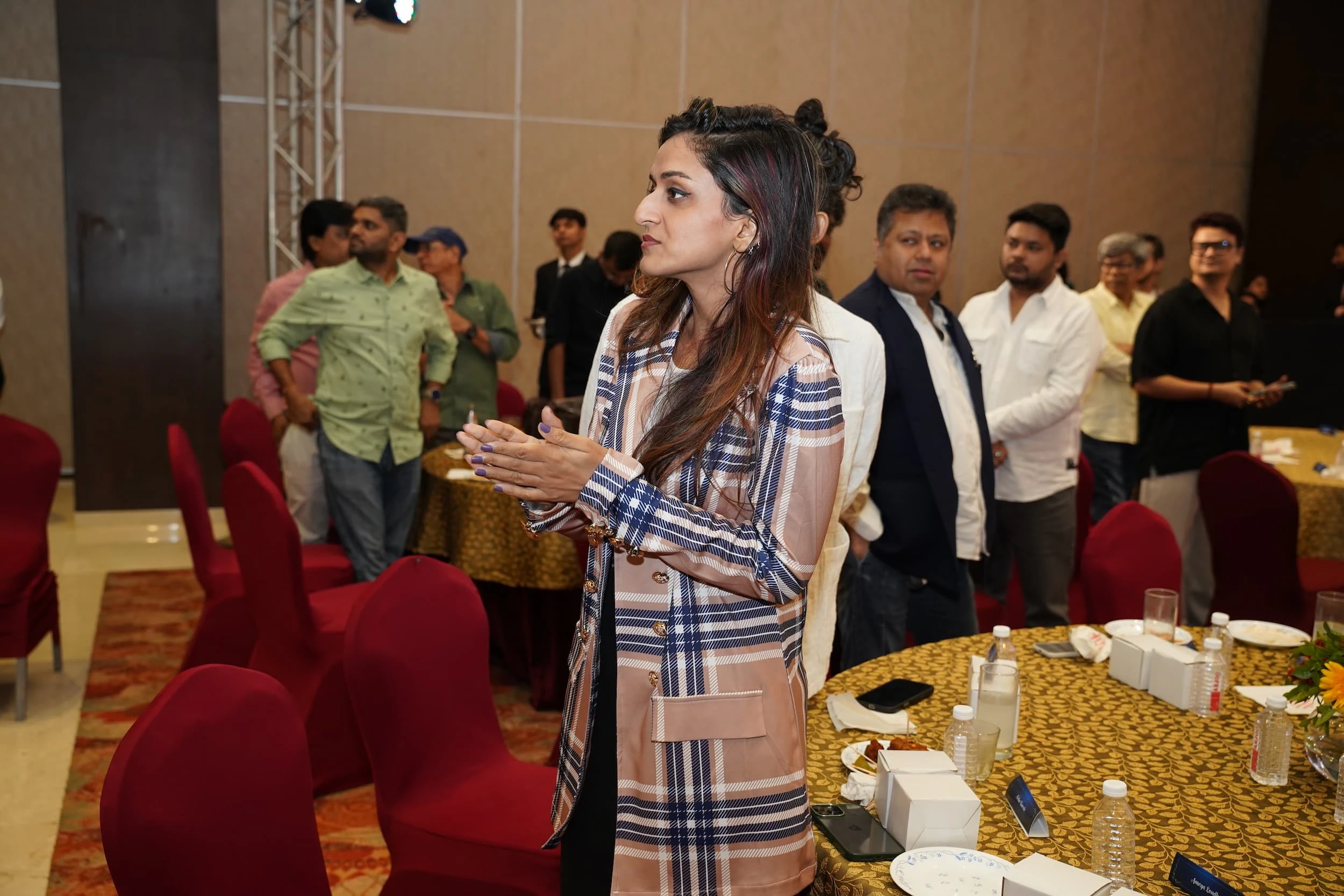 Woman in plaid outfit at indoor event with attendees standing around tables.