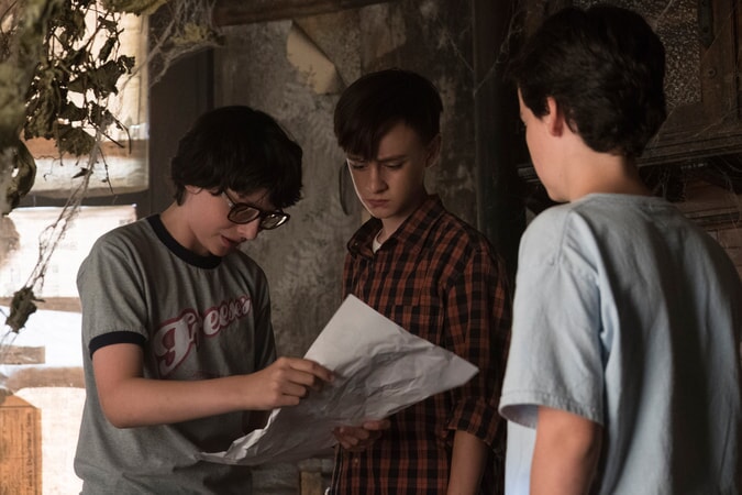 Three of the kids from the IT movie stand looking at a piece of paper
