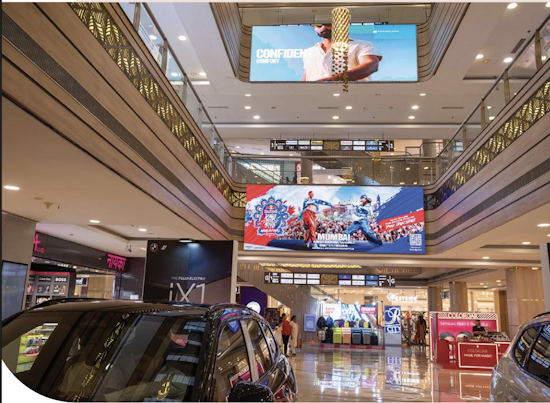 Shopping mall interior viewed from the ground floor, showing upper levels, displayed cars, retail stores, and multiple digital screens
