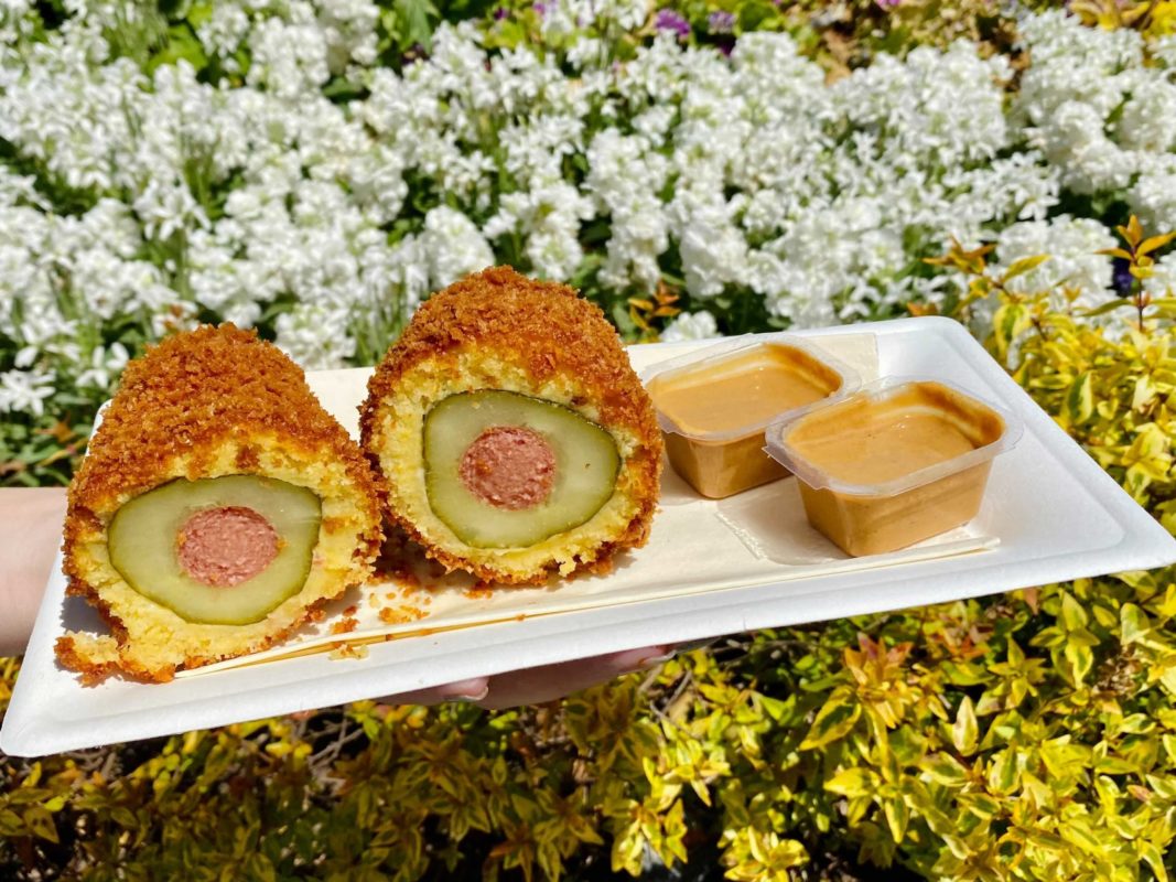 In front of a white flower bush, there is a Styrofoam plate with a corn dog cut in half with 2 cups of peanut butter. The corn dog is cut in half to reveal a hot dog inside of a pickle, dipped in fried pancake batter