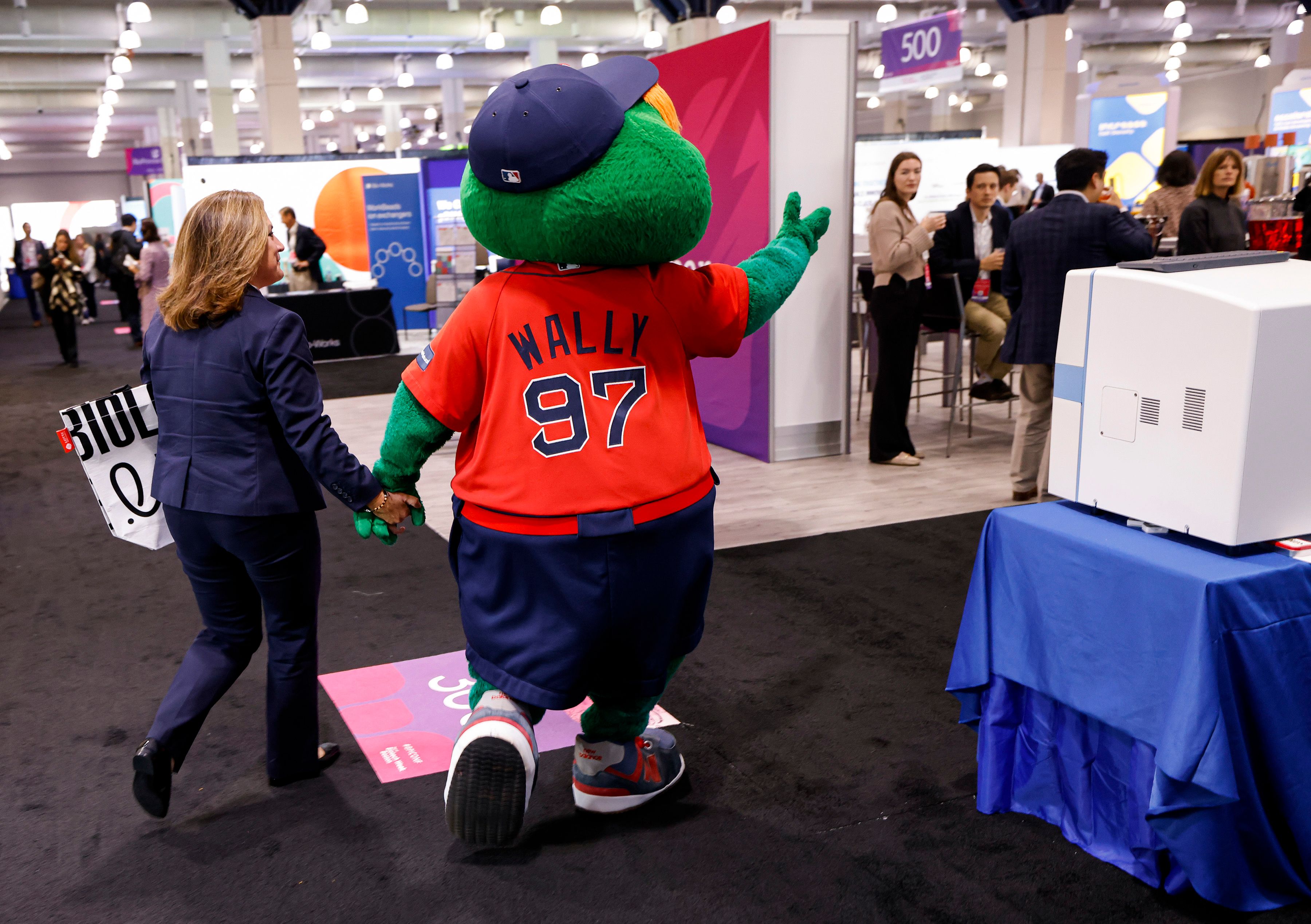 Wally the Green Monster mascot in an orange t-shirt, walks hand in hand with a female bioprocessing professional in the exhibit hall at BioTech Week Boston.
