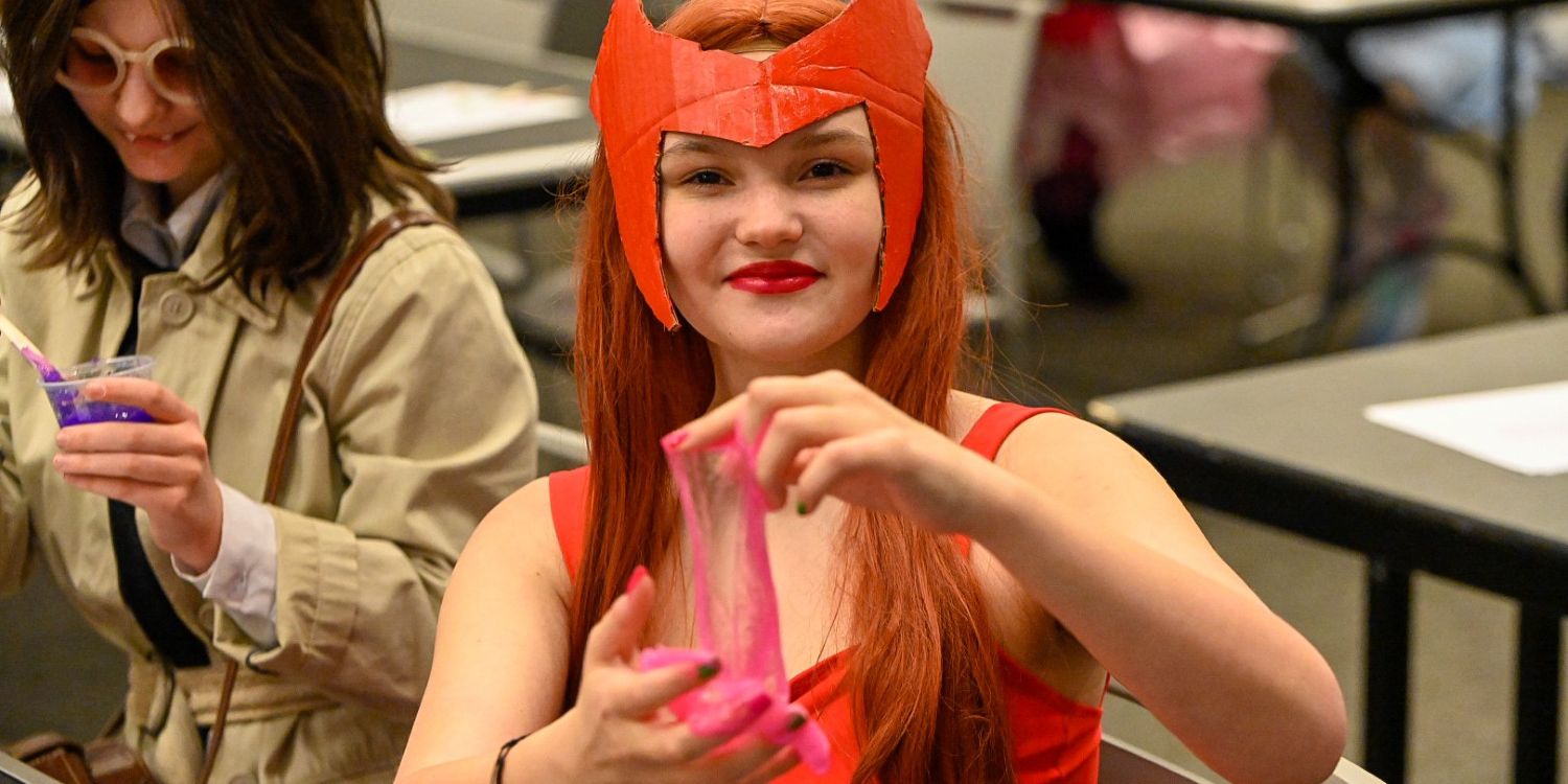 A young girl dressed as a Ghostbuster holds up a cup of pink slime, smiling at the camera. Another attendee in a trench coat and round sunglasses stirs a cup of purple slime in the background.
