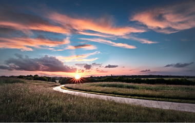 landscape photo of a grassy field with a one-way road winding through it. Sun is setting behind a hill and the sky is blue with a few pink clouds in the sky.