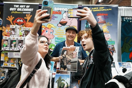 The image captures a candid moment at a convention booth, where a person is taking a selfie with a smartphone, and another individual is holding up a signed illustration, looking excited. In the background, Jeff Brennan is smiling at the camera, standing behind a display of colorful artwork. The environment suggests a fan or comic book convention, where fans interact with artists and collect autographs or artwork. The expressions and activity in the photo convey a lively and engaging atmosphere typical of such events, where attendees celebrate and support creative work.