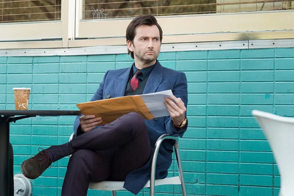 Wearing a blue suit, David Tennant is sitting in a metal chair in front of a teal tile wall as he holds a manila file and pulling out sheets of paper