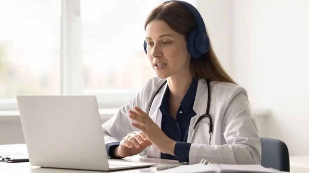 Médica usando jaleco e fones de ouvido realizando atendimento de telemedicina em notebook sobre a jornada híbrida na saúde.