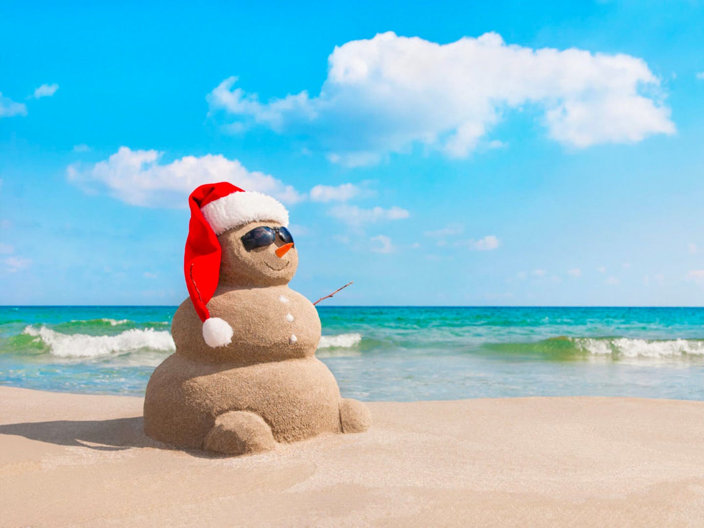 A snowman made of sand sits on a beach with a red santa hat on