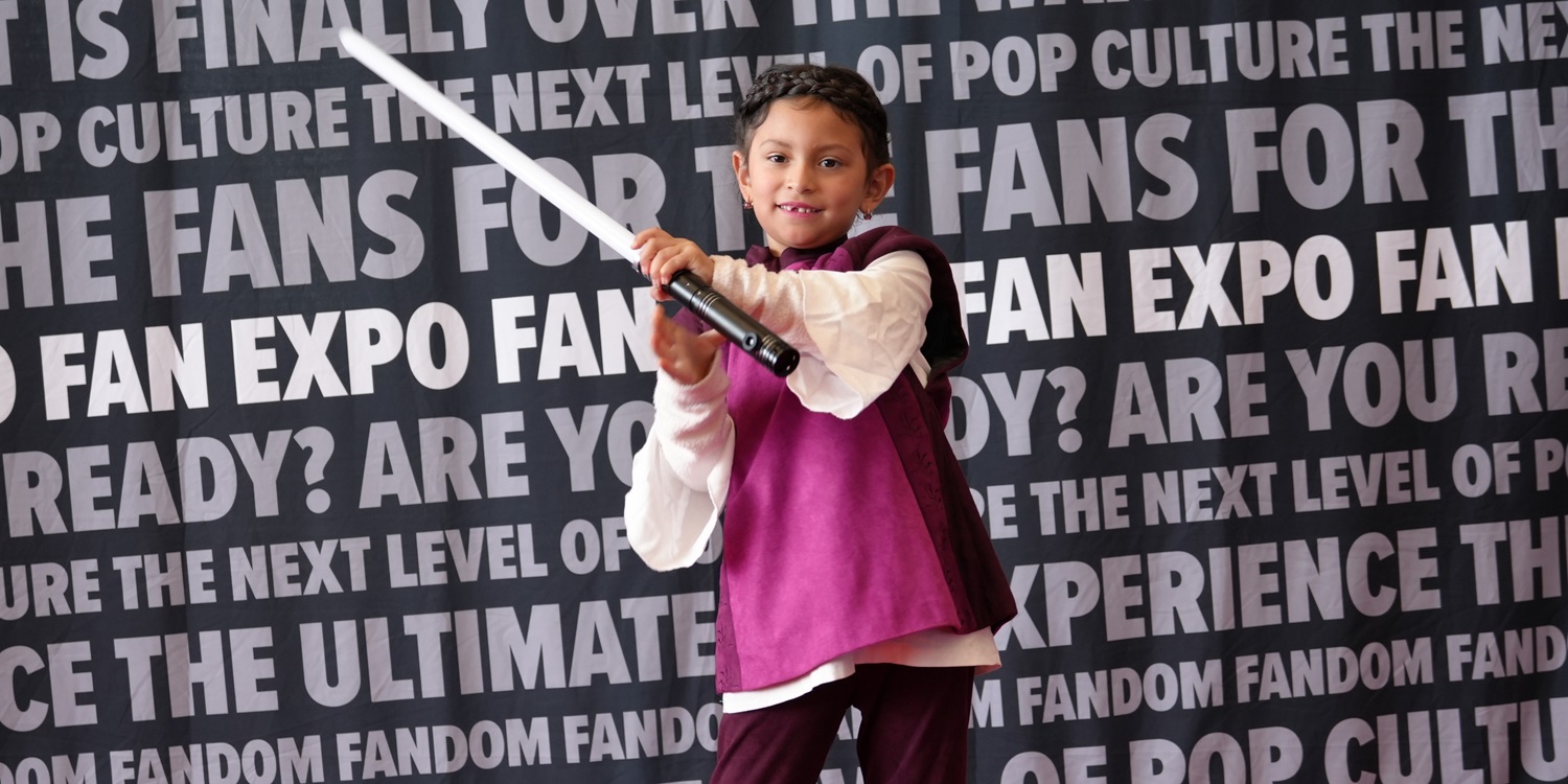 A young child dressed in a fantasy-style costume with a purple vest and white sleeves wields a toy lightsaber, smiling confidently. Behind them, a FAN EXPO-themed backdrop with bold, capitalized text promotes pop culture and fandom excitement.