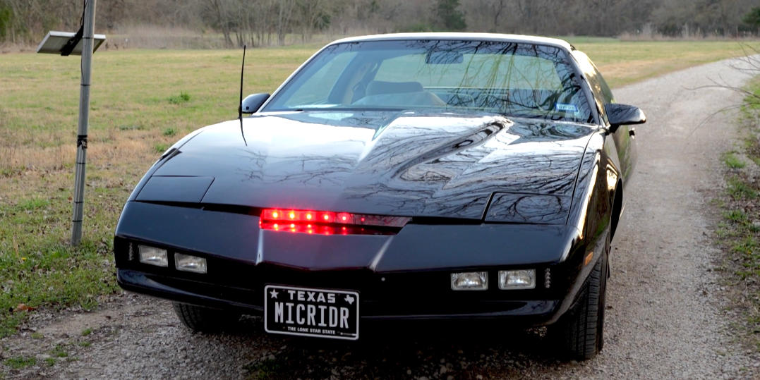 Front view of the DFW Knight Rider replica car with its signature red scanner lights glowing, parked on a gravel path with fields and trees in the background.
