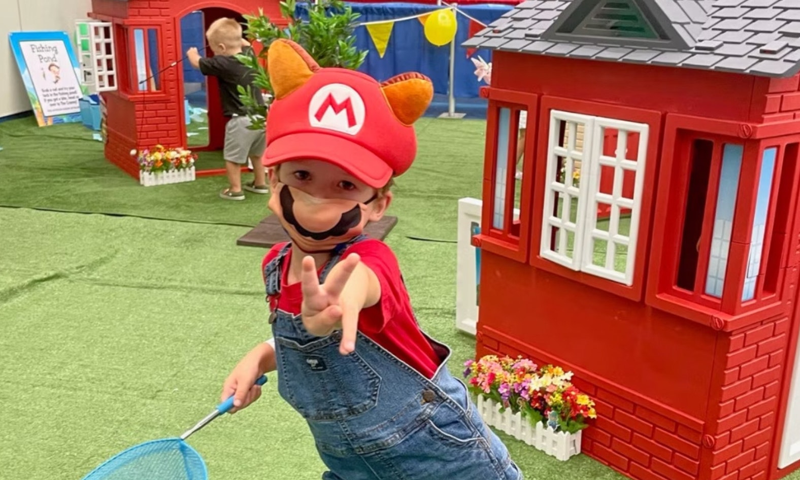 A young boy poses in a Mario cosplay. He is wearing a red Mario hat with ears, blue overalls and a red t-shirt. He is holding a blue butterfly catcher in one hand.